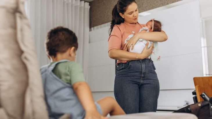 A mother lovingly cradles her baby while her child sits nearby on the couch in a modern room setting.